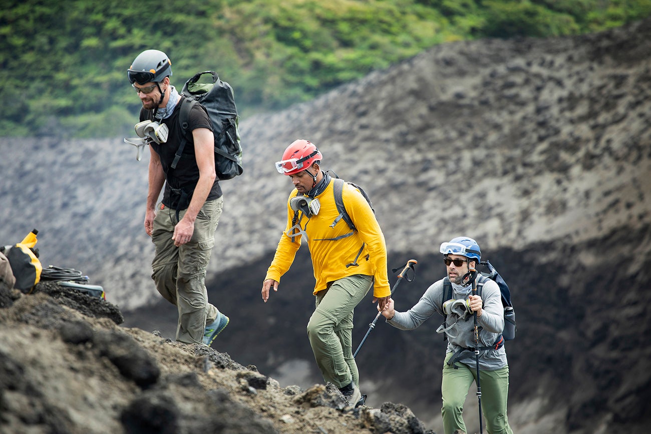 Three men climb out of an active a volcano wearing helmets, and respirators around their necks. The third man, Eric, walks with two guiding poles.