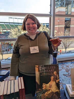 Ann Campbell standing at a table at the American Society of 18th Century Studies event with her book titled "Families of the Heart: Surrogate Relations in the Eighteenth-Century British Novel" over a painting of two adults and a child dressed in 18th-century clothing