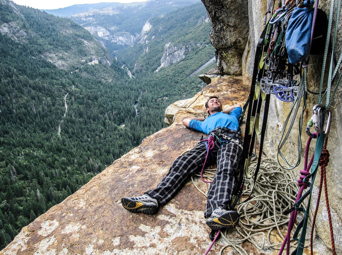 Dane Larson relaxes with his climbing gear on a rock shelf on the side of a mountain, a valley sprawling out behind him.
