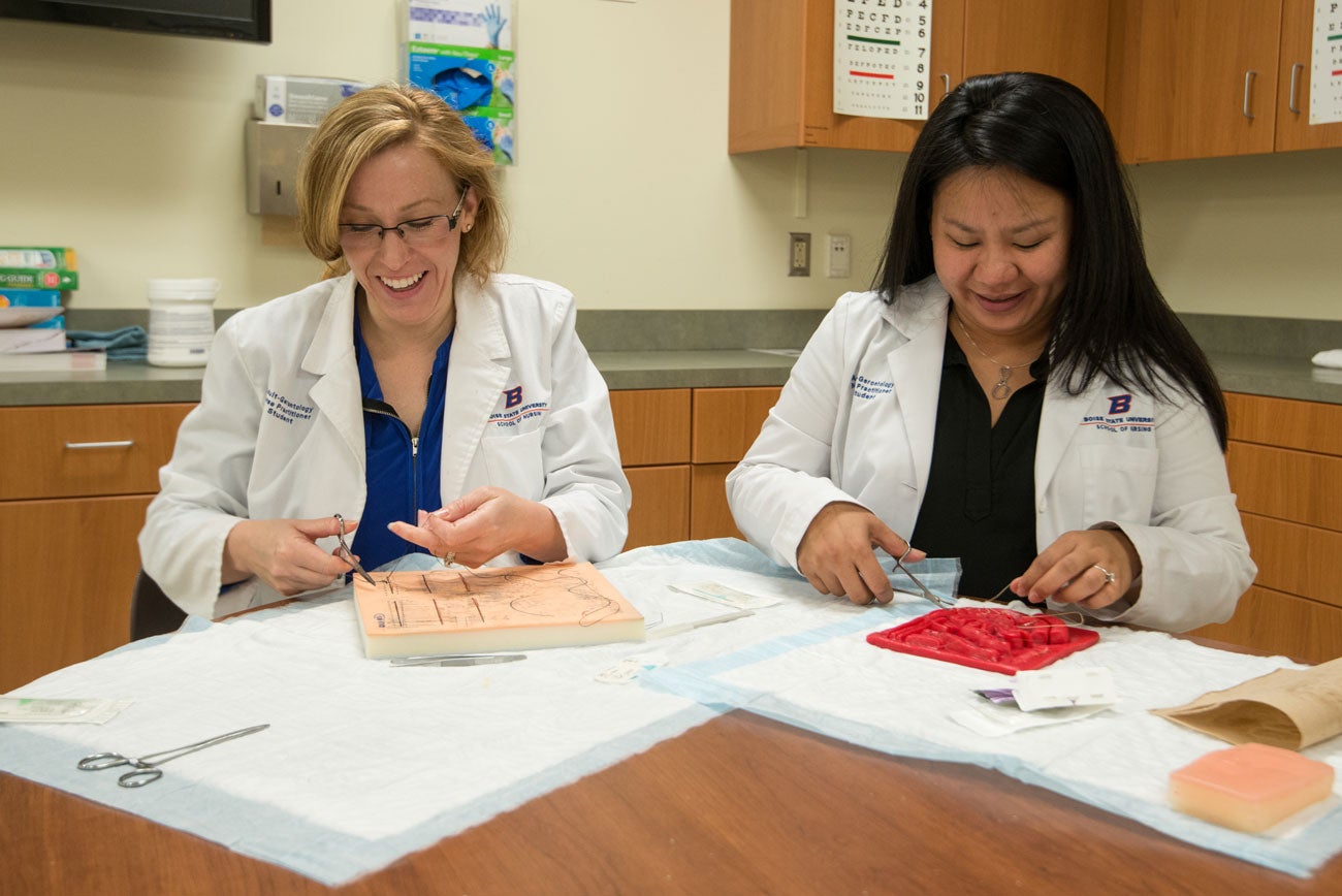 Two nurse practitioner students sit behind a table and practice suturing.