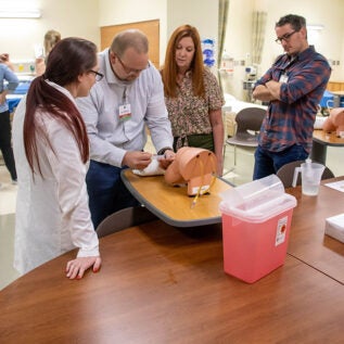 A nurse practitioner student practices giving a joint injection while their professor looks on.