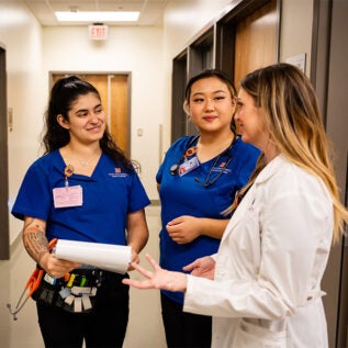 A nurse in a white coat talks to two smiling student nurses in blue scrubs.