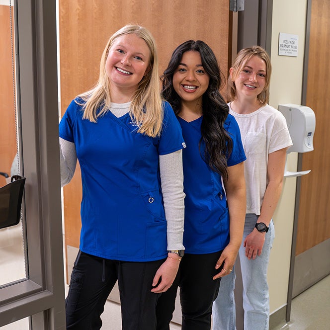 Three nursing students stand in the doorway of a simulation center room.