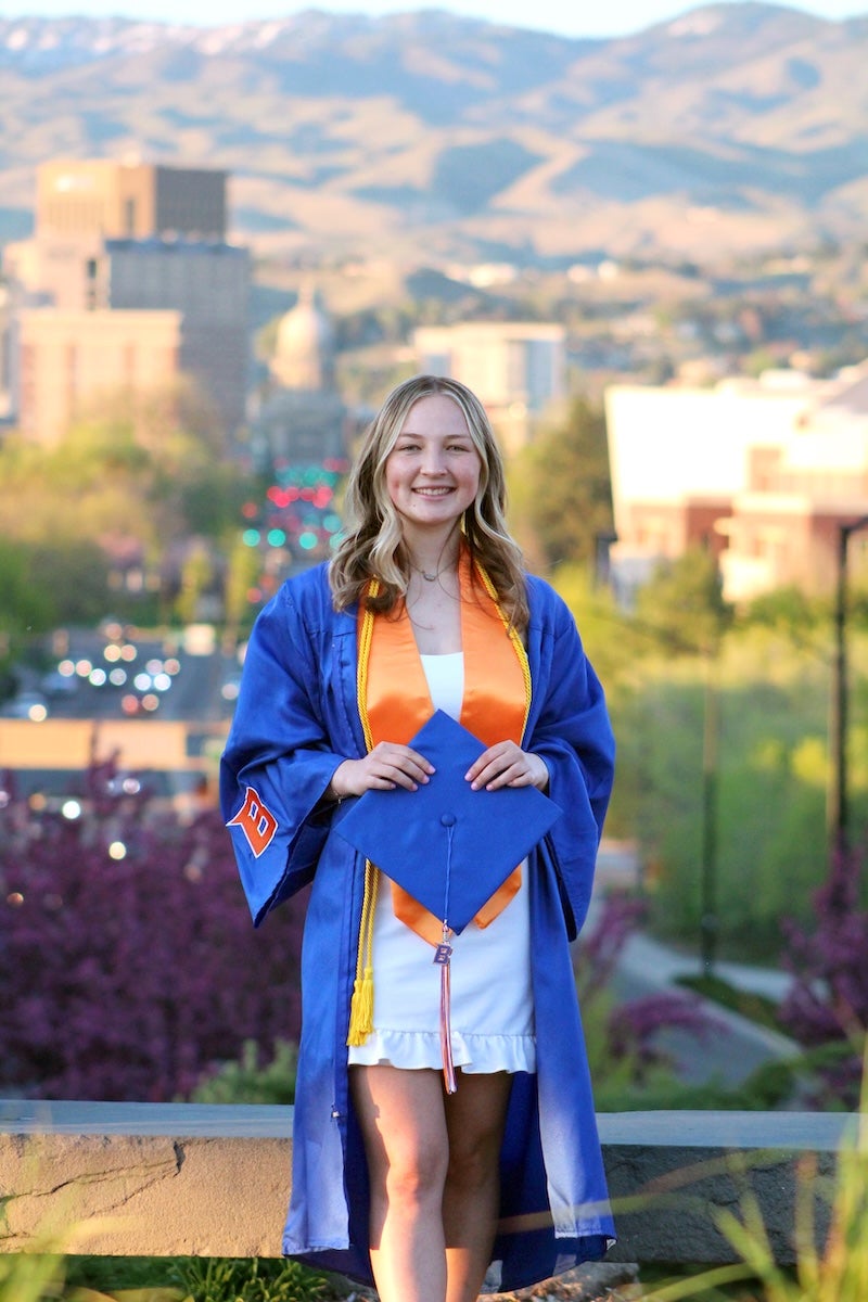 A person in grad regalia stands at the Boise Depot with downtown in the background