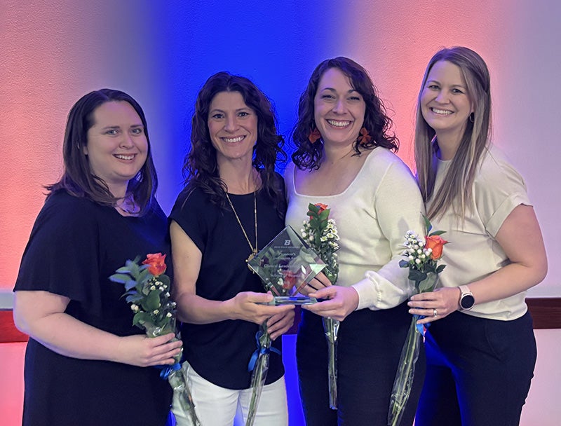Four women hold roses and a plaque while standing in front of a wall lit up orange and blue.