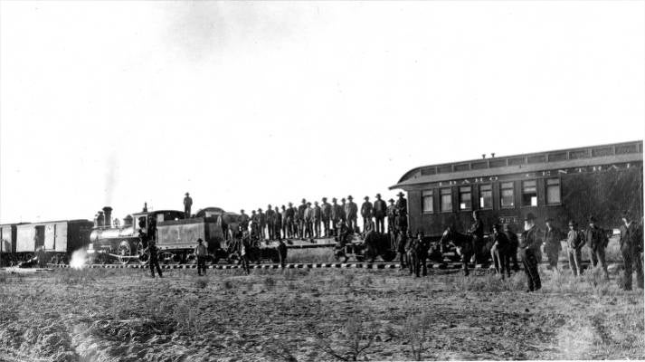 black and white photograph shows group of men standing on a flat railway car, steam engine to the left.