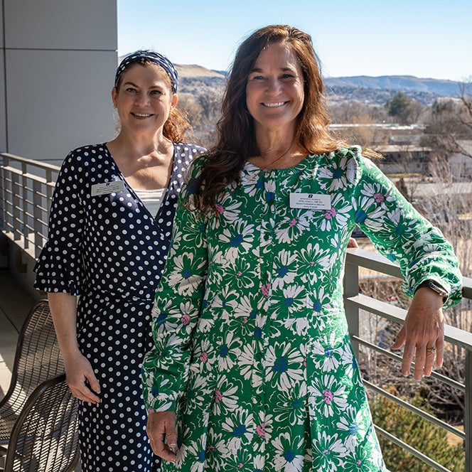 Two women in floral dresses smile and stand on a balcony with the Treasure Valley behind them.