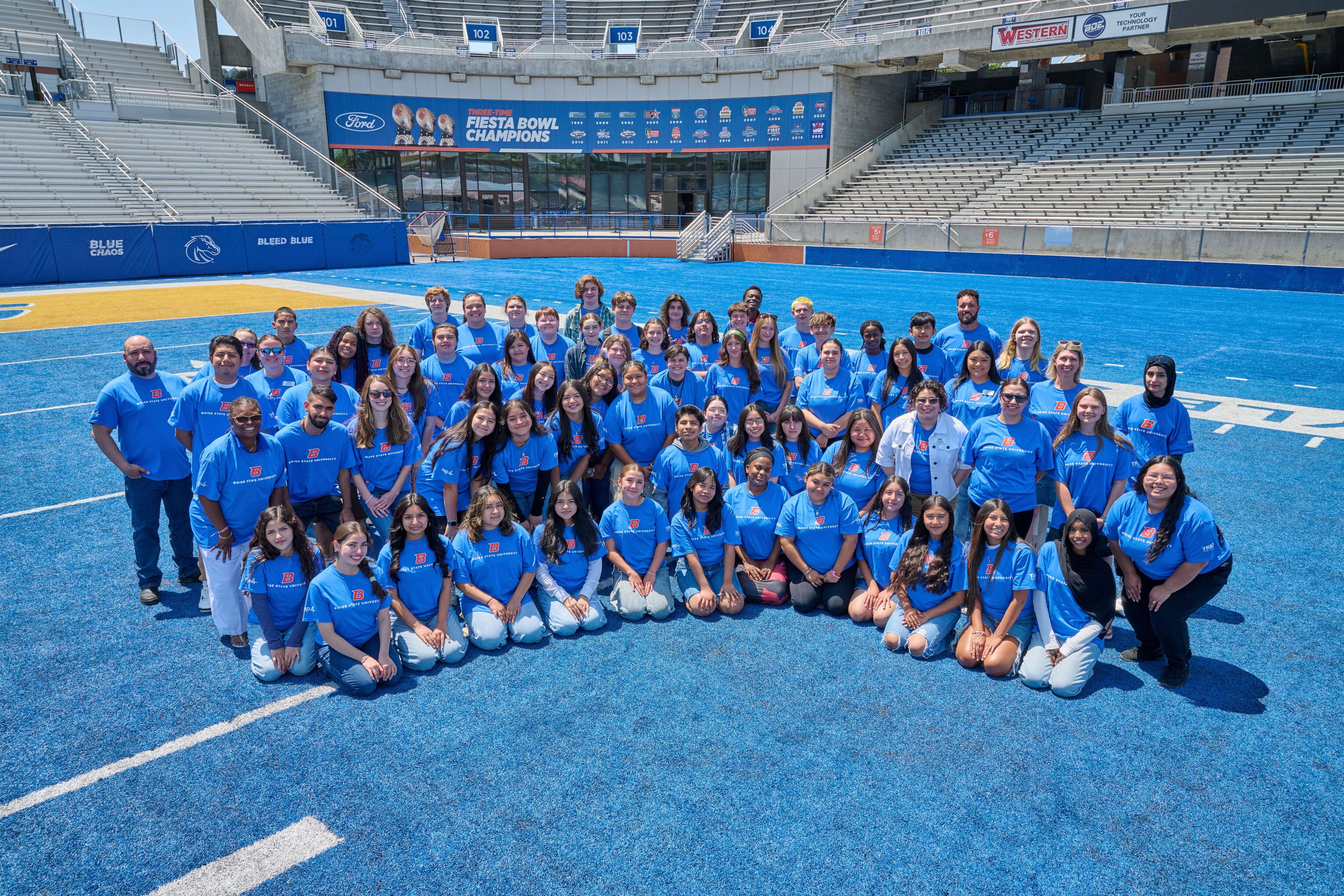 A group of REP4 students, all wearing blue t-shirts, pose with Boise State faculty and staff on the famous blue turf