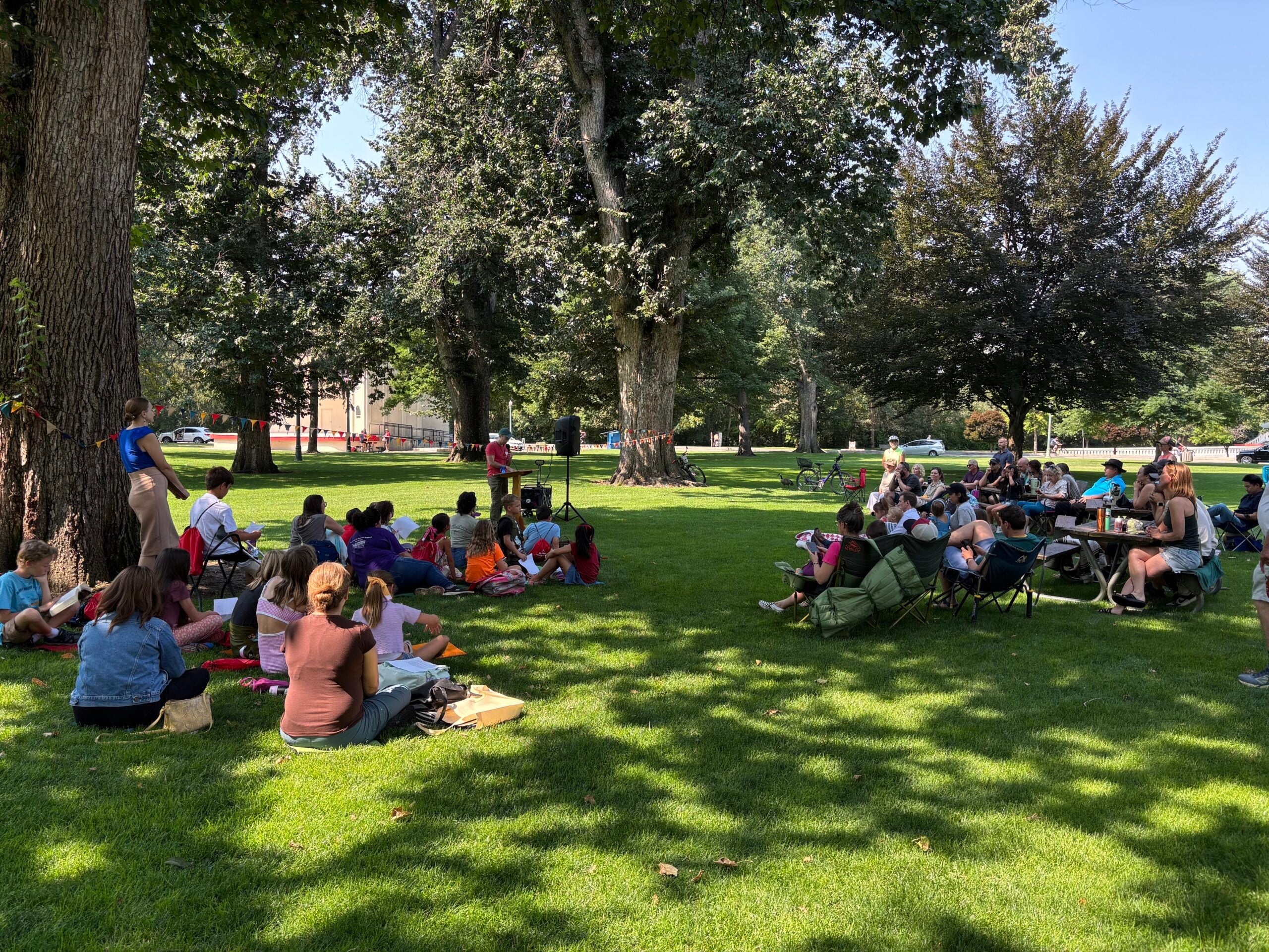 A group of people sit in the grass and listen to a speaker