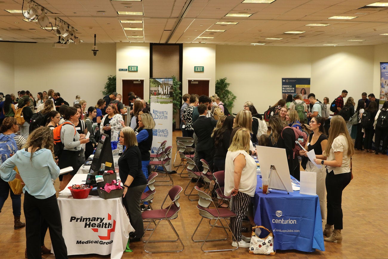 People mingle around two rows of tables set up for a nursing job fair.