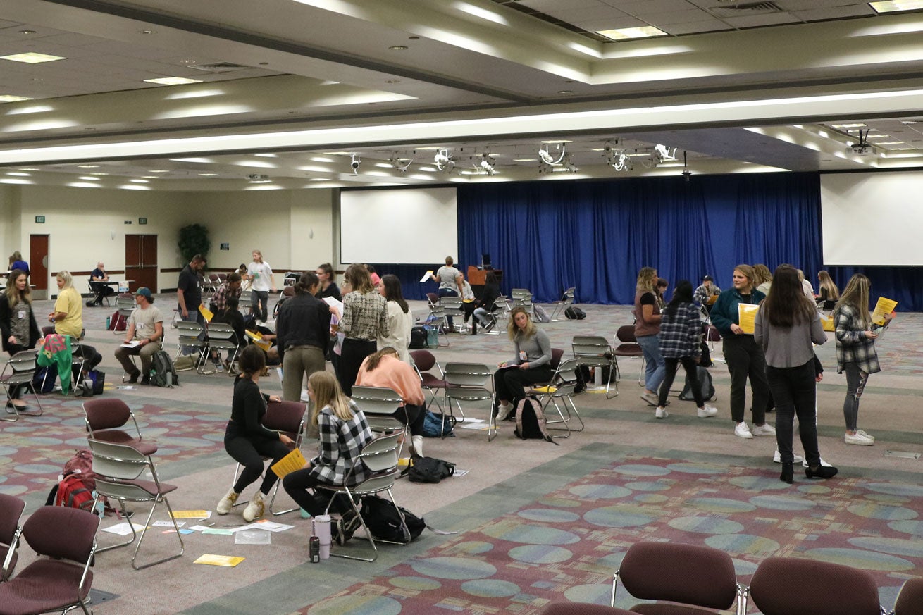 Students mill about groups of chairs in a ballroom during the Poverty Simulation.