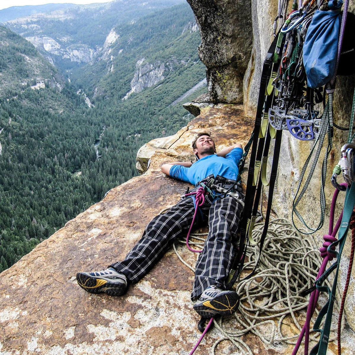 Dane Larson relaxes with his climbing gear on a rock shelf on the side of a mountain, a valley sprawling out behind him.