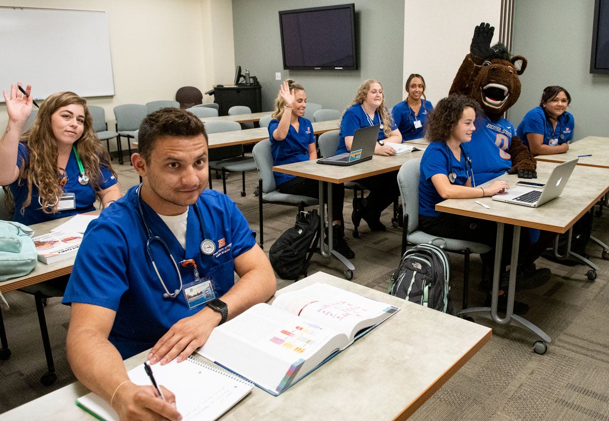 A classroom is full of nursing students raising their hands. Buster Bronco sits at a table among them.