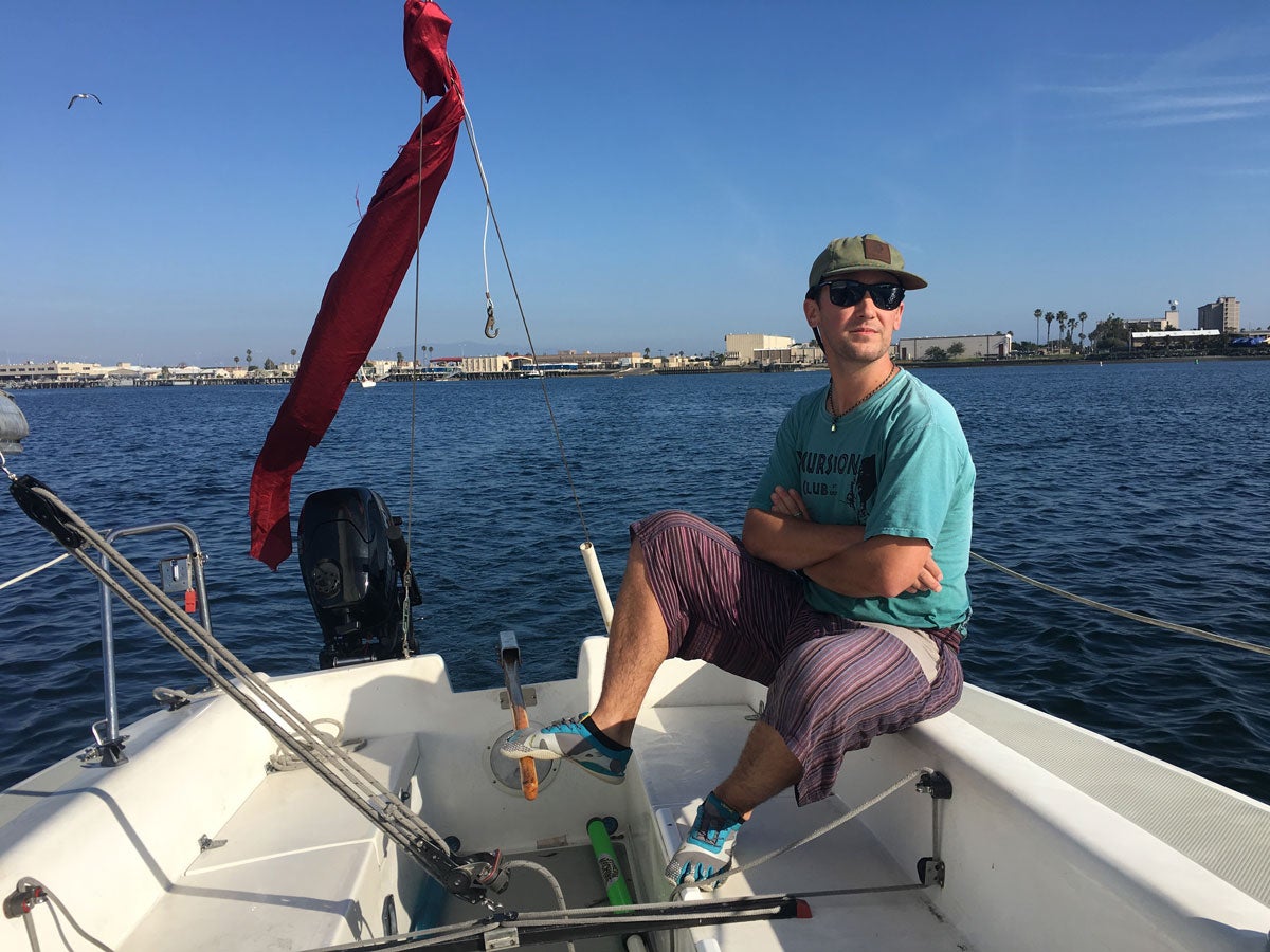 Dane Larson sits by the stern of a boat in the middle of a bay, palm trees and the city behind him.