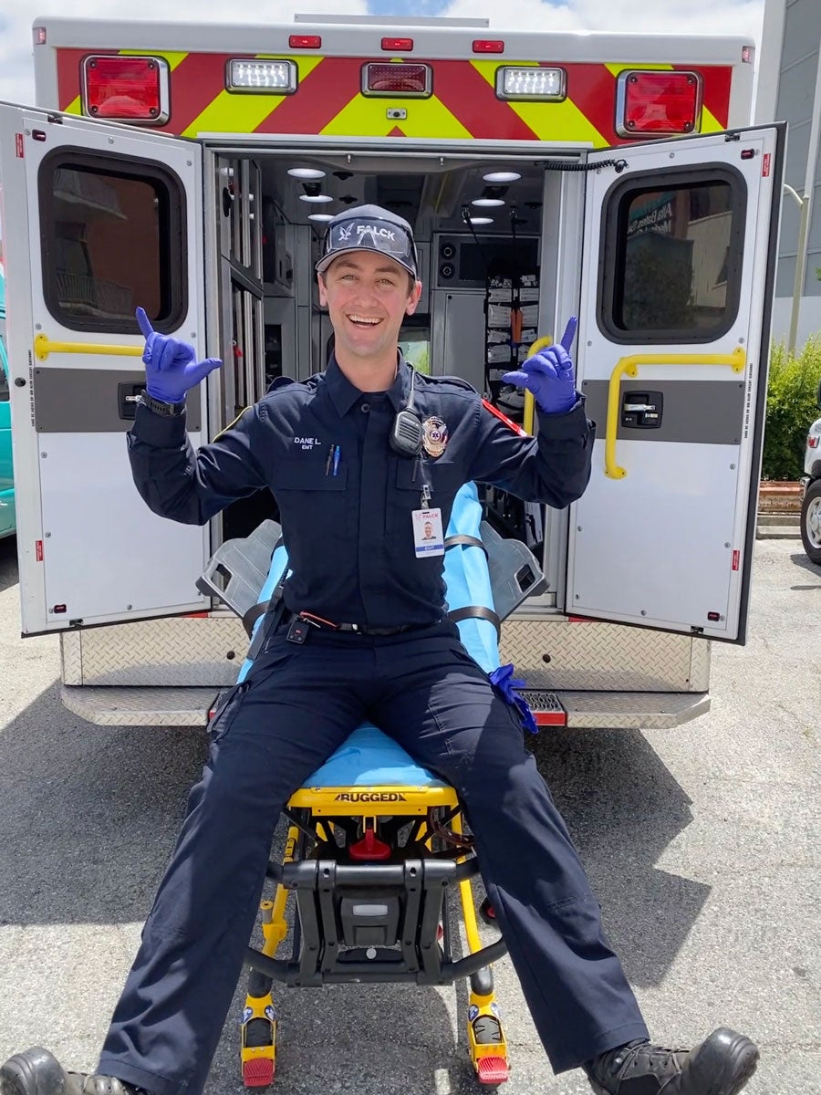 Dane Larson wears his EMT uniform and smiles, sitting on a gurney out the back of an ambulance. 