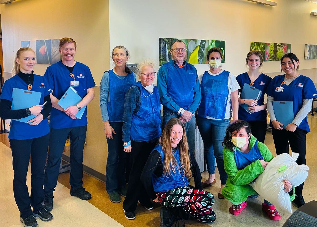 Nursing students in scrubs stand with standardized patients who portray patients in a behavioral health setting, distinguished by blue pinnies.