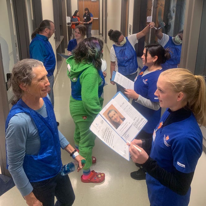 Students in blue scrubs hold open folders and conduct behavioral health assessments with standardized patients in a hallway.