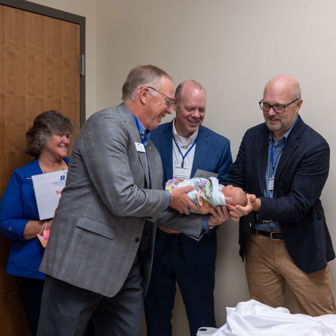 Three men in suits smile as they pass off a manikin baby to one another.