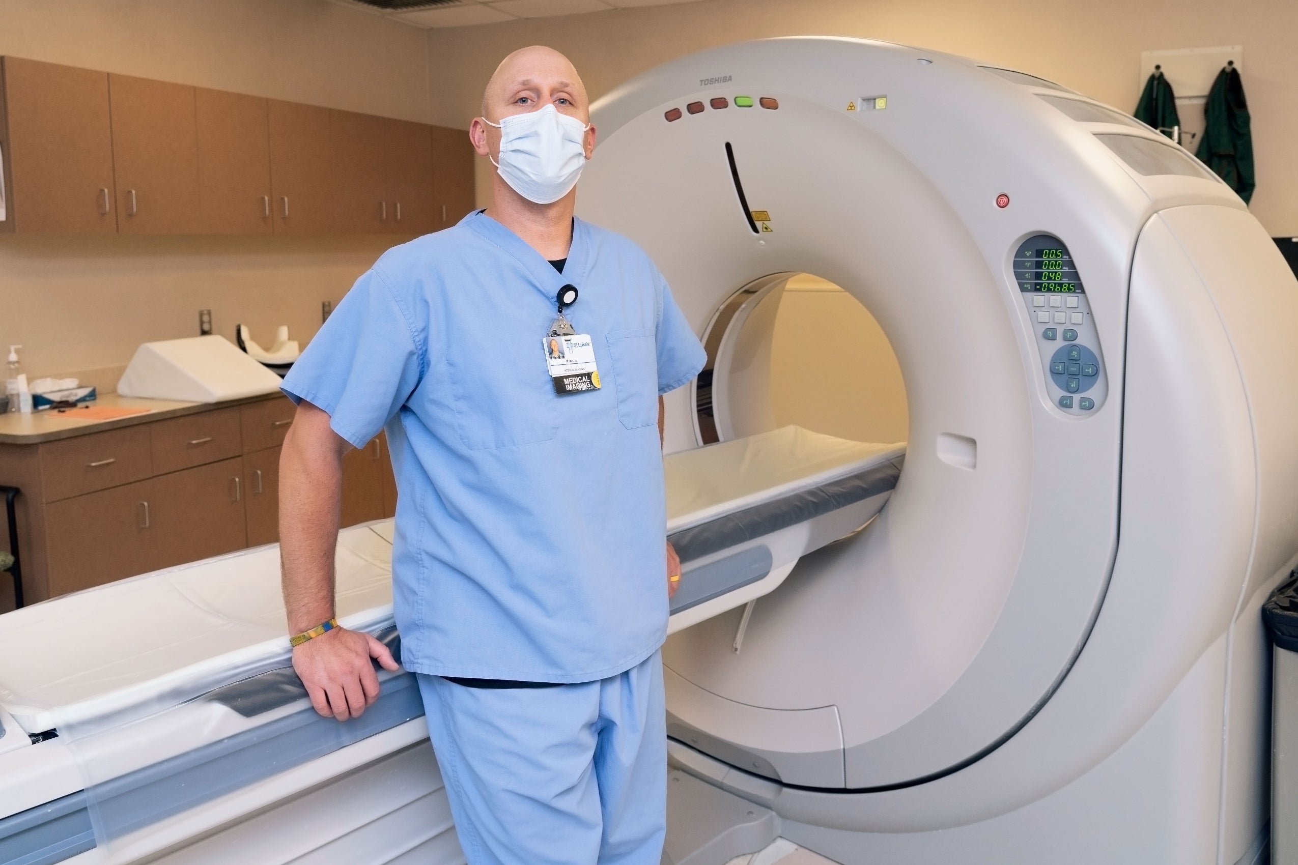 Imaging Sciences Student in blue scrubs, standing in front of a CT Scanner.
