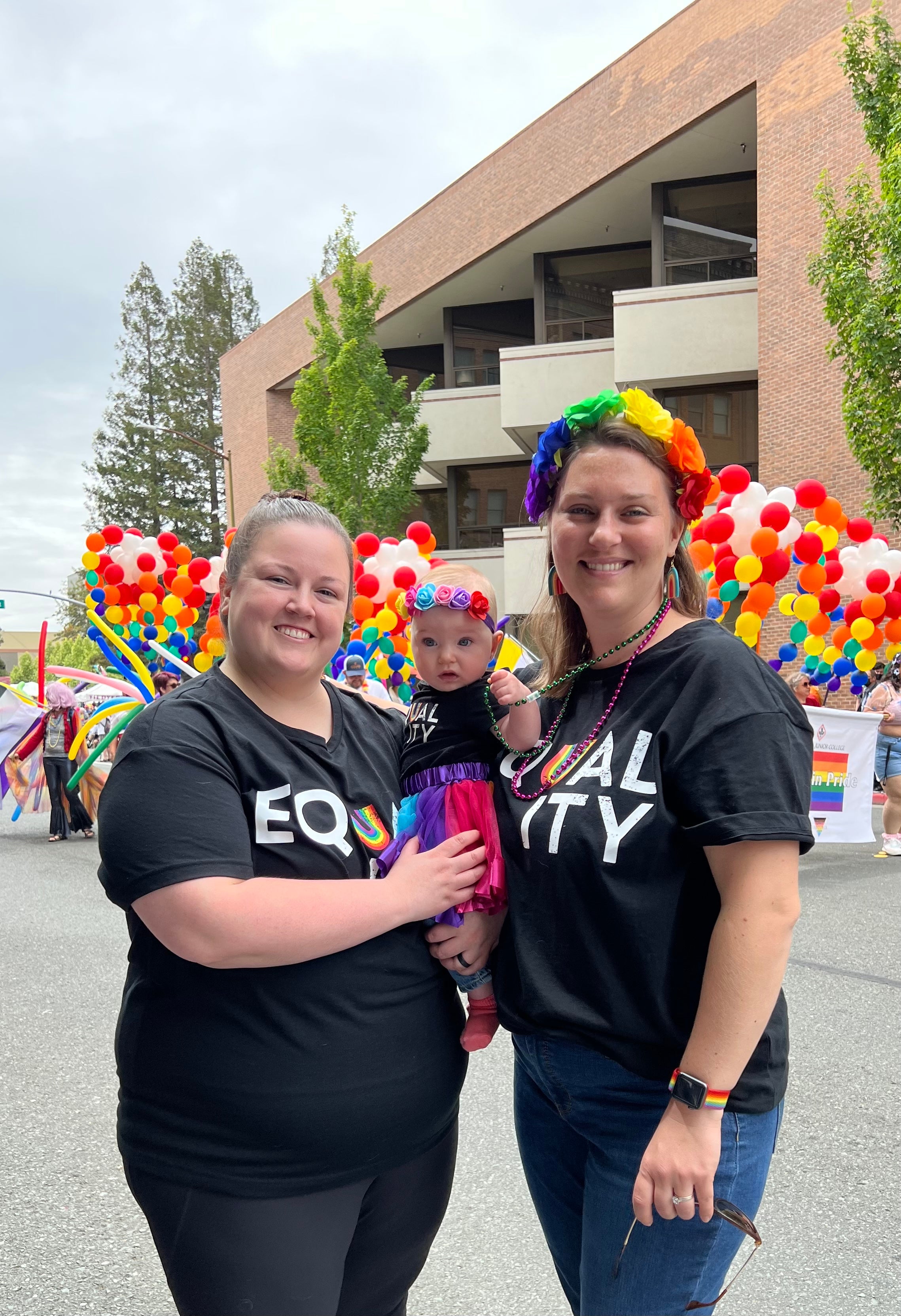 Kim stands next to another woman holding a baby, rainbow balloons and banners are in the background