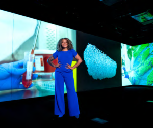 woman wearing a blue jumpsuit stands before vibrant, interactive display