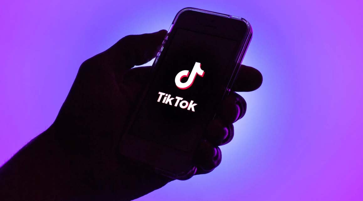 A sillohuette of a hand against a purple background, holding a phone with the TikTok logo on the screen