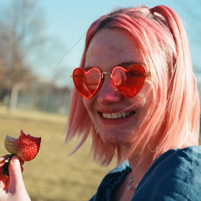 Naomi smiles at the camera and holds a strawberry. She is wearing heart shaped sunglasses.