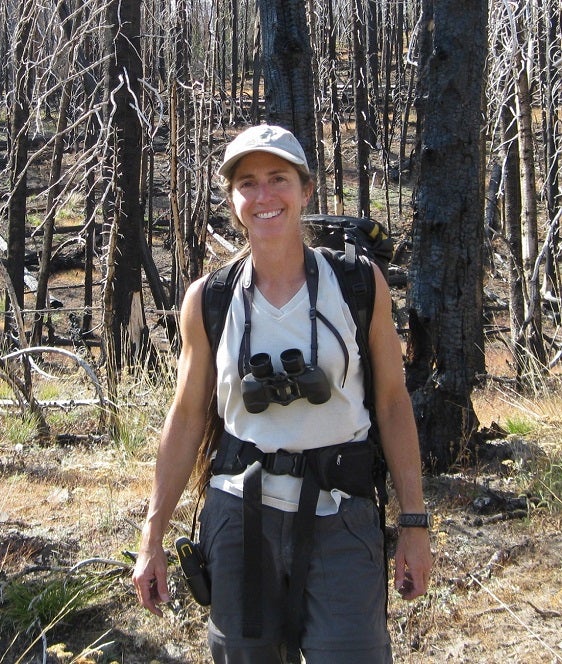 Alumna Robin Garwood standing among burned whitebark pine trees with binoculars