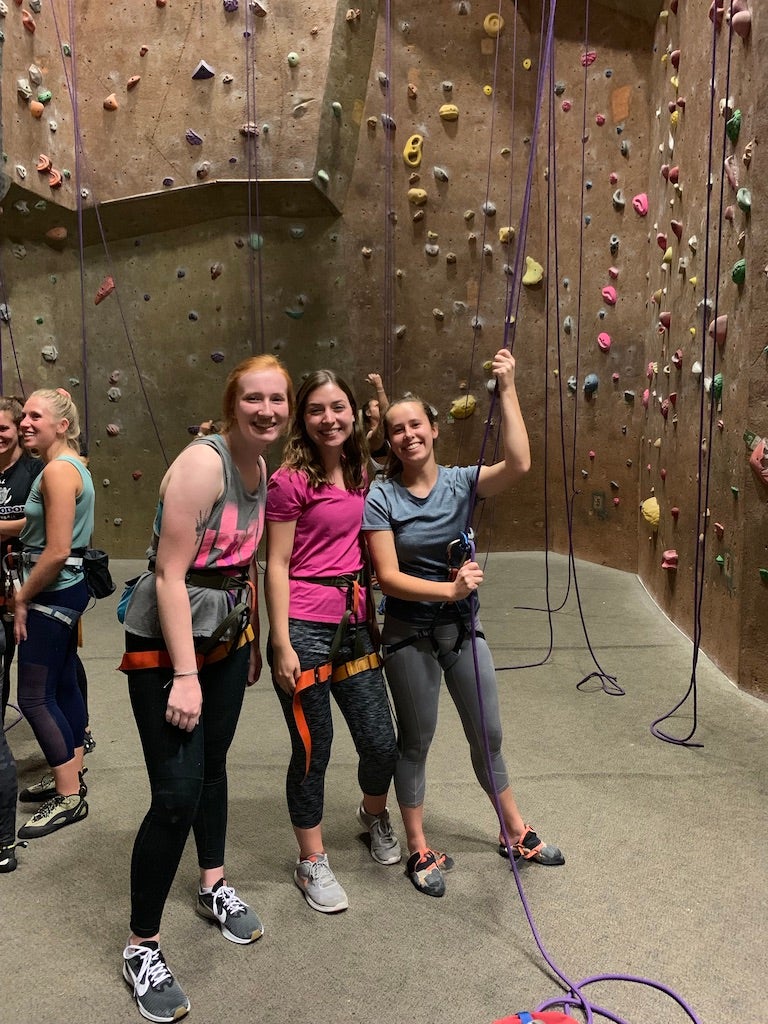 3 students in climbing gear stand next to climbing wall