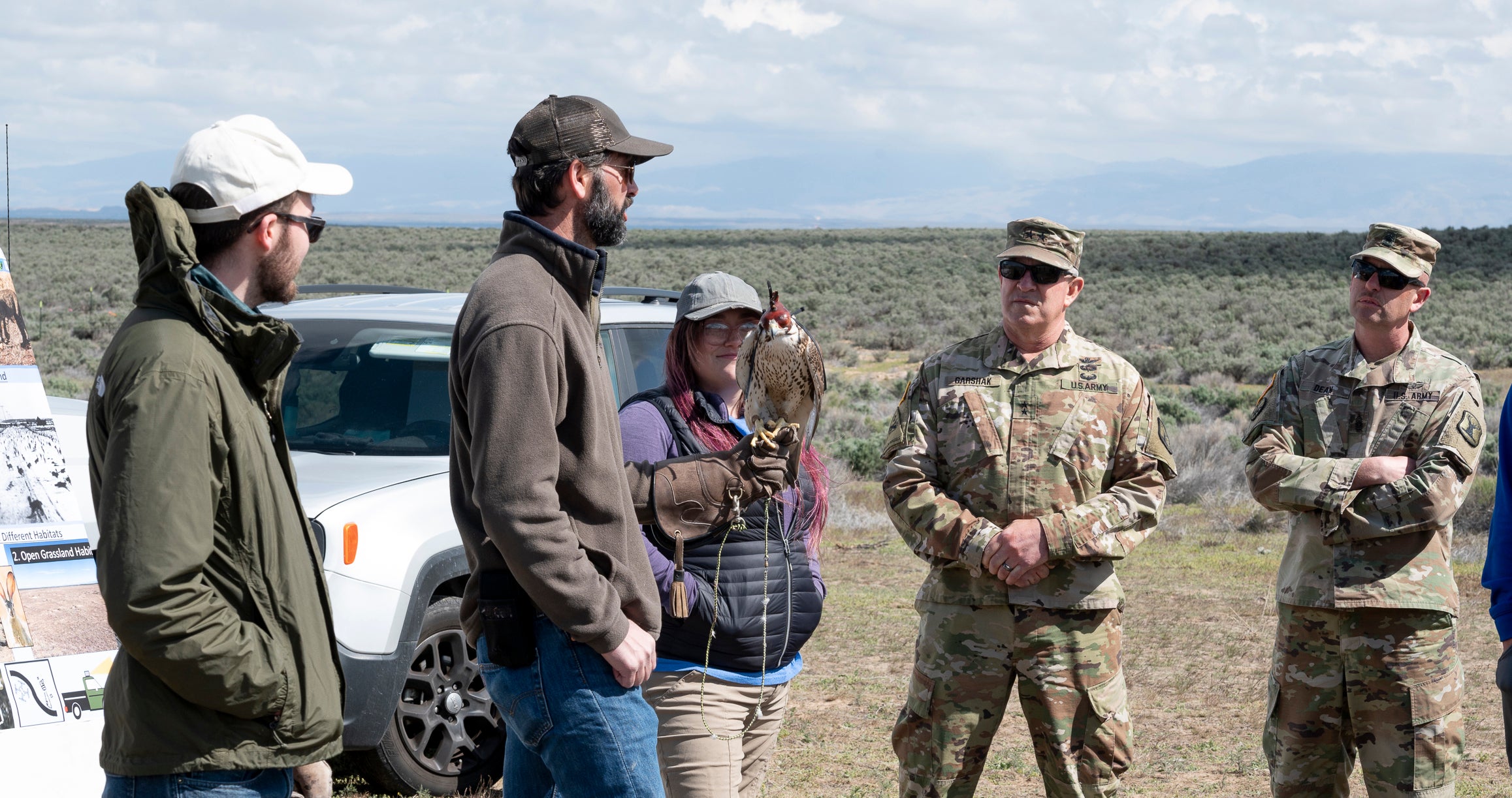two armed forces memebrs and three bioligists stand outside in sagebrush steppe. one man holds a hooded raptor
