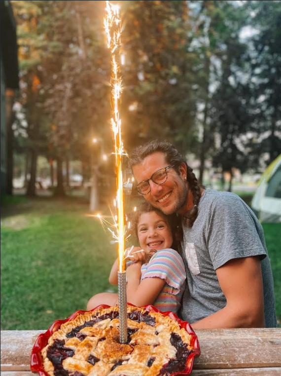 A picture of Adam Musser and his daughter Josie sitting at a picnic table with a delicious birthday pie with a firework in it!