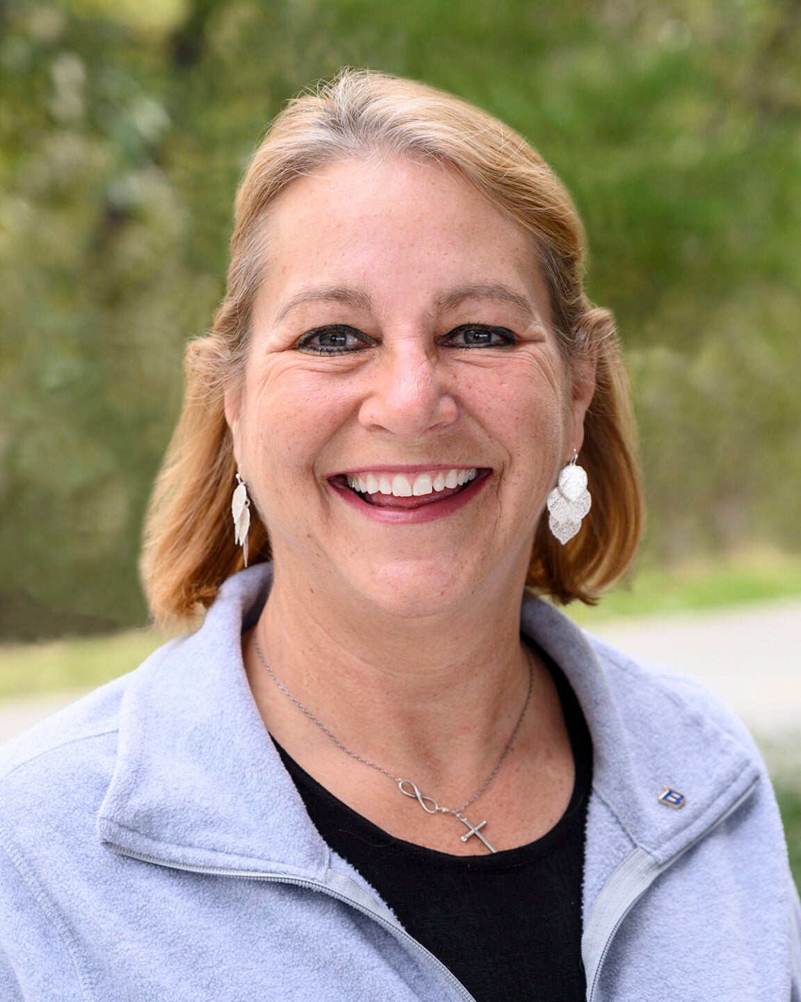 A photo of Dana Oster, a middle-aged woman with shoulder-length blonde hair, smiling in an outdoor setting.