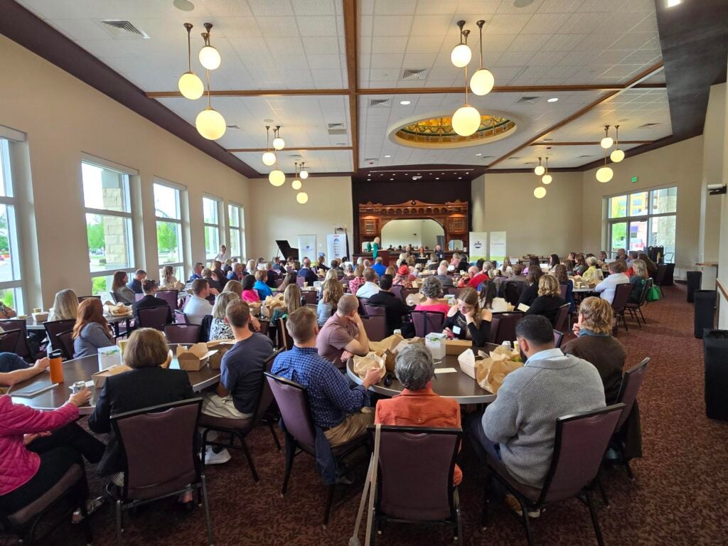 Many people sit at round tables, listening to the City Club panel.