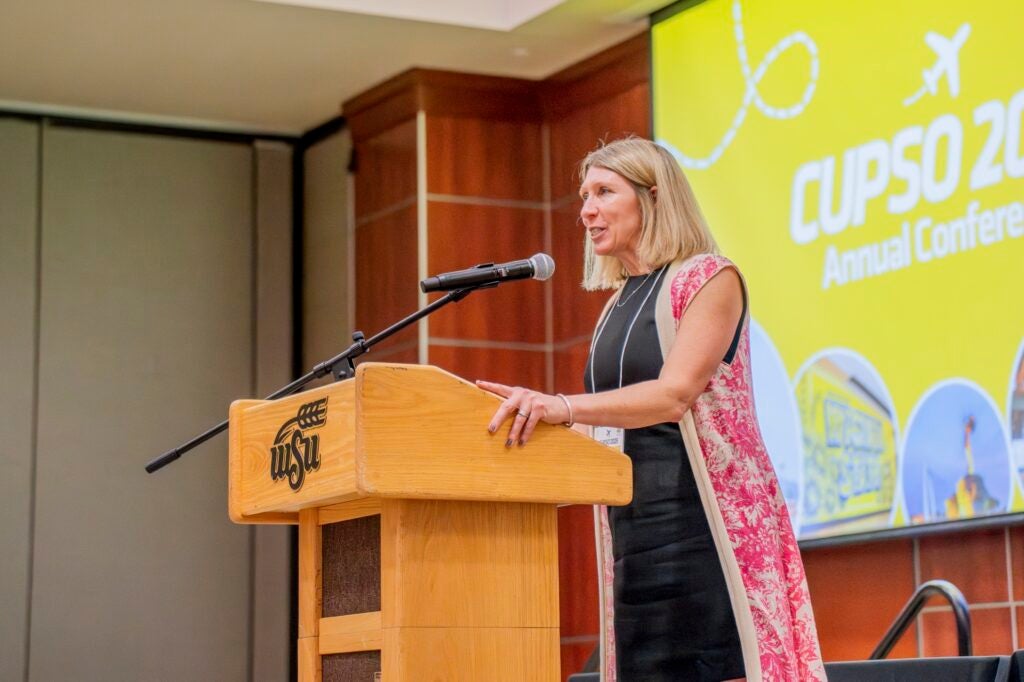 A woman stands behind a lectern, in front of a screen with the CUPSO logo.