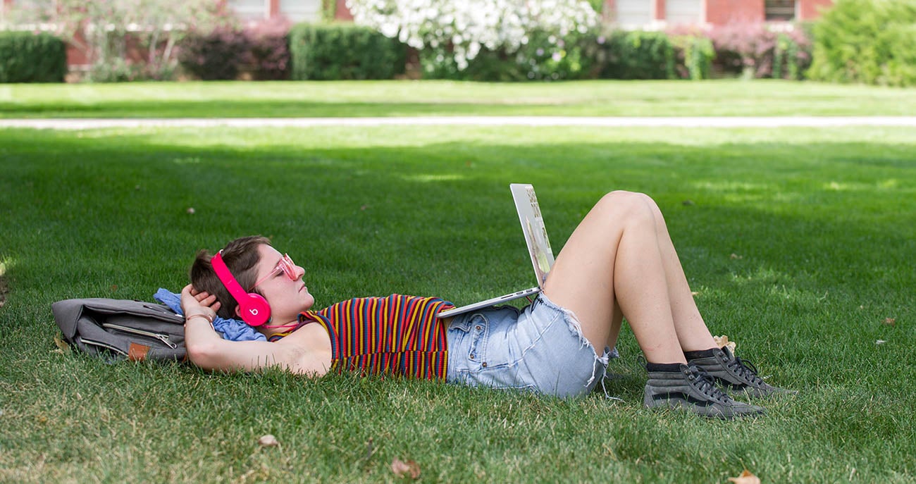 Student lying on the grass, with headphones on, looking at a laptop