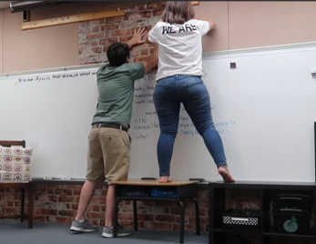 Two teachers hanging up something on a wall with one of the teachers using a desk and cabinet as a ladder