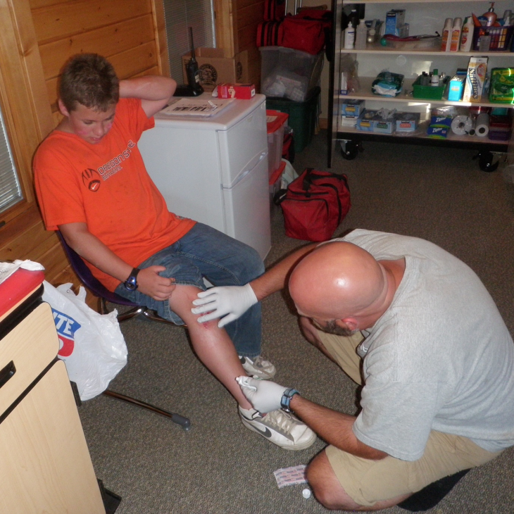 A volunteer nurse kneels in a cabin to apply ointment to a cut on the knee of a boy in an orange shirt.