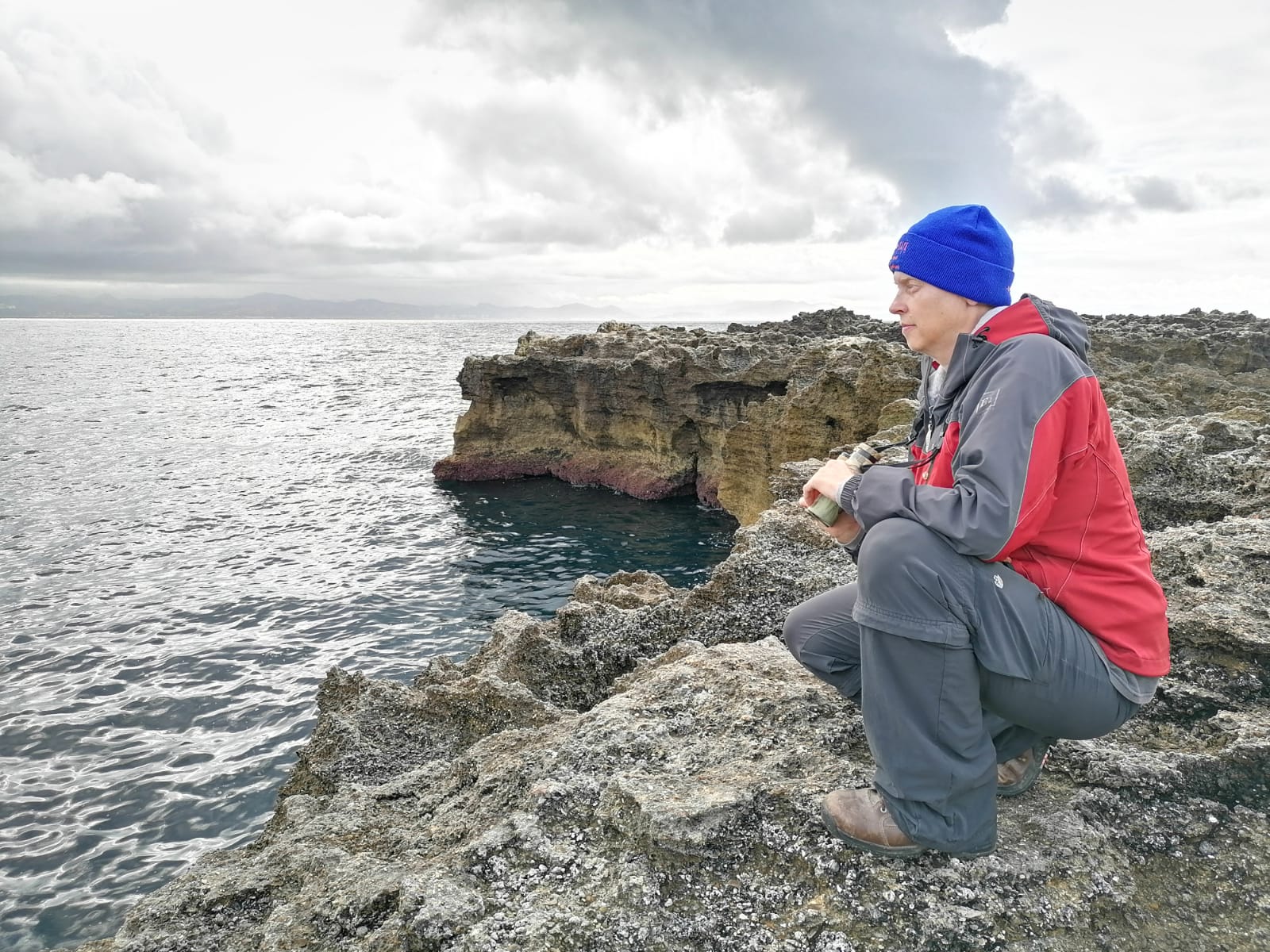 a scientist in a boise state hat kneels on a cliff edge overlooking the ocean, holding binoculars