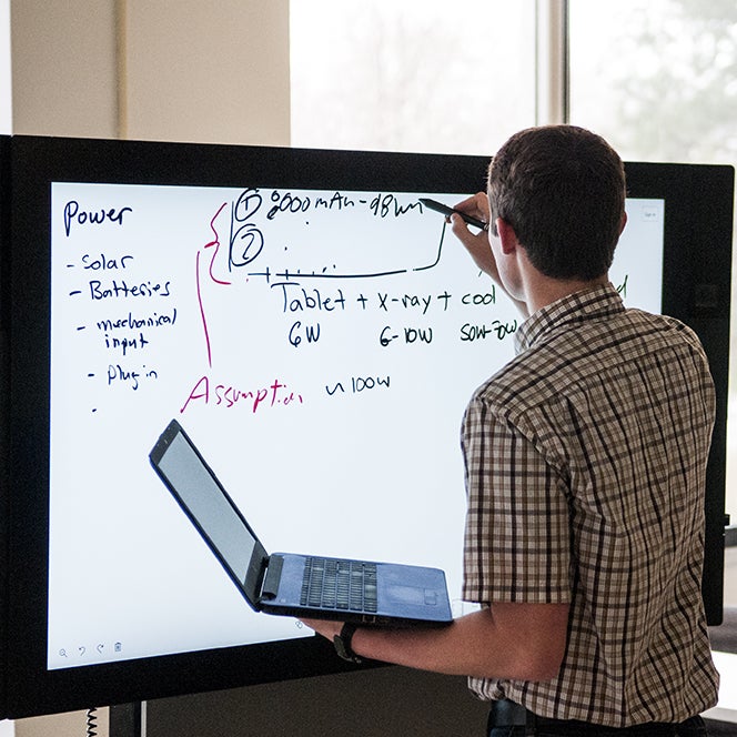 Students working on whiteboard while holding laptop