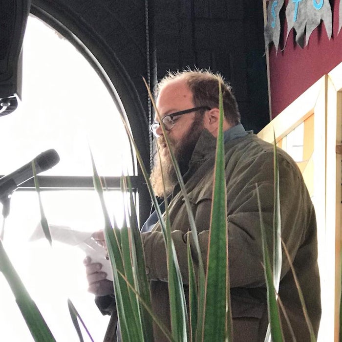 The author at a reading. A bright window is in the background and plants in the foreground. 