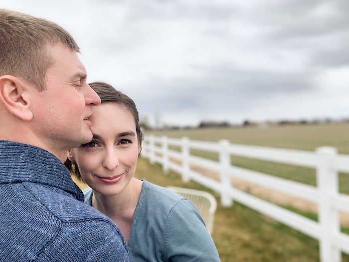 The author leans against her husband. A cloudy sky, a white fence, and fields are in the distance.