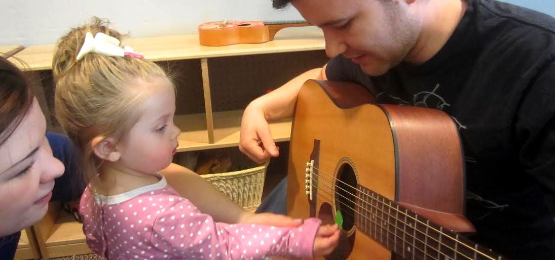 Male staff holding guitar while toddler girl strums its strings with the help of her mother