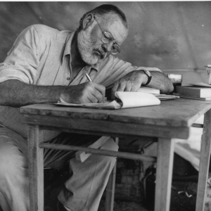 A black and white photo of Ernest Hemingway sitting in a chair, leaning slightly over a table and writing in on a notepad.